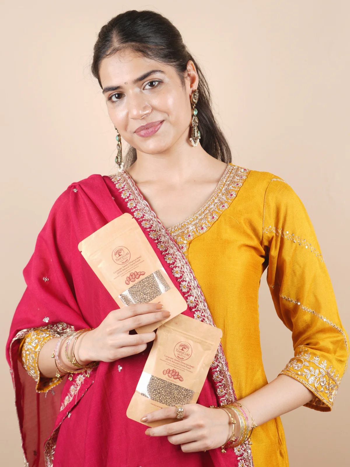 Smiling woman in a festive suit holding two packs of Taaru Gold Coriander Seeds